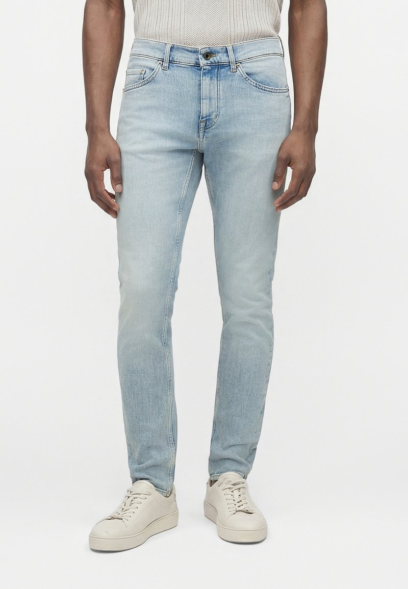 Άνδρας που φοράει στενά ανοιχτά μπλε τζιν με κούμπωμα κουμπιού και φερμουάρ, συνδυασμένα με λευκά δερμάτινα αθλητικά παπούτσια σε μονόχρωμο φόντο.