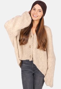 Cardigan en tricot beige avec des manches oversize et un ourlet court, présentant des gros boutons à l'avant. Porté avec un jean foncé et un bandeau marron.