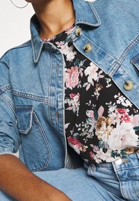 Denimjakke i en lys blå nuance, med to frontlommer og guldknapper, lagdelt over en sort blomstret top i blødt stof.