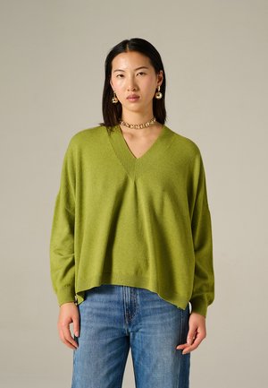 Maglione verde lavorato a maglia con scollo a V, vestibilità ampia e orli a coste, abbinato a jeans azzurri a gamba dritta. Texture piatta e aspetto morbido.