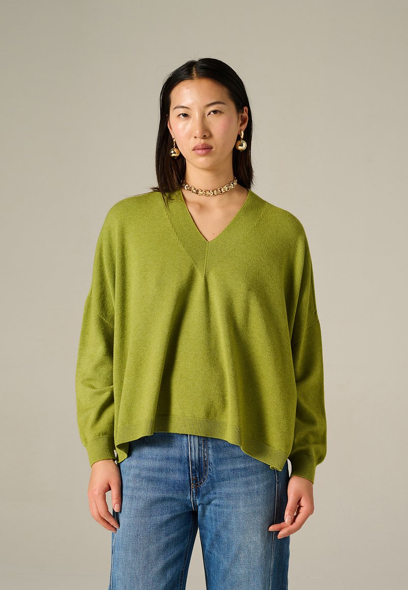 Maglione verde lavorato a maglia con scollo a V, vestibilità ampia e orli a coste, abbinato a jeans azzurri a gamba dritta. Texture piatta e aspetto morbido.
