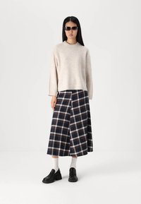 Μπεζ πλεκτό πουλόβερ, πλισέ καρό φούστα σε μπλε, λευκό και κόκκινο. Μαύρες παπούτσια με τετράγωνο τακούνι; γυαλιά ηλίου; κάλτσες ορατές. Μινιμαλιστικό φόντο.