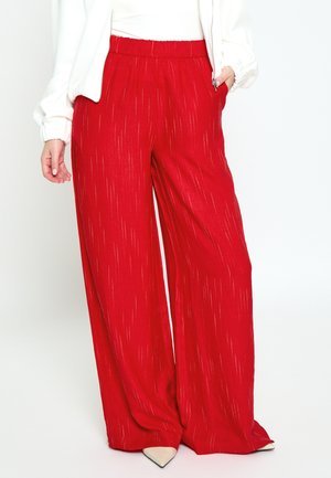 Žena nosí široké červené nohavice s jemnými vertikálnymi pruhmi, v kombinácii s bielymi špicatými topánkami a bielym dlhým rukávom.