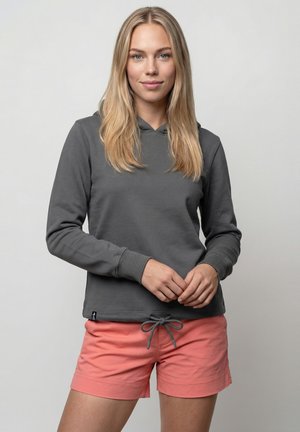 Bl blonde Frau trägt einen dunkelgrauen Kapuzenpullover und korallrote Shorts mit Kordelzug, steht mit verschränkten Händen vor einem schlichten hellen Hintergrund.