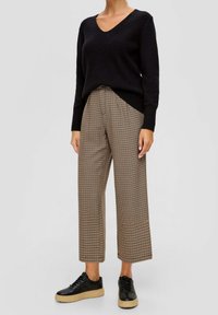Pull noir à côtes avec col en V, associé à un pantalon large à motif à carreaux beige et marron, complété par des baskets noires à semelle épaisse.