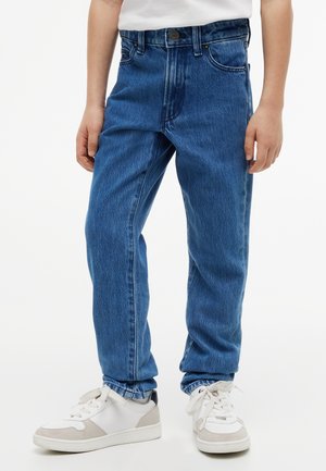 Enfant portant un jean en denim bleu et des baskets blanches avec des touches beiges, debout devant un fond uni.