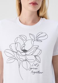 Balta marškinėliai su juodu eskizu atvaizduojančiu magnolijos žiedą ir užrašu "Augkime kartu" po juo.