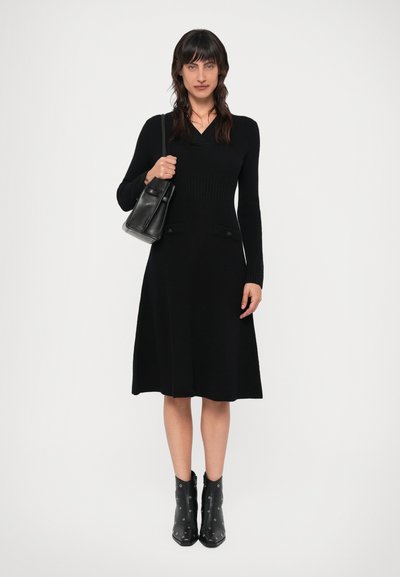 Musta neulemekko V-pääntiellä, pitkät hihat ja ribbikuvio; siinä on kaksi sivutaskua. Yhdistetty mustiin niitattuihin nilkkureihin ja pieneen laukkuun.