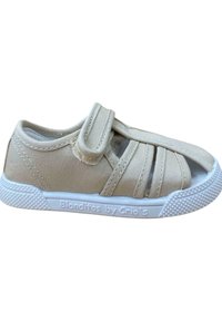 Zapatilla de lona beige con suela de goma blanca y correa de velcro. Presenta un diseño troquelado y una suela con patrón texturizado. Etiqueta de la marca en el lateral.