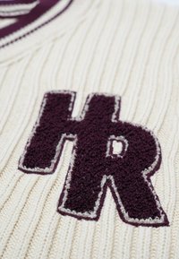 Krämfärgad ribbad stickad tröja med en strukturerad mörkröd "HR"-logotyp med vit kontur, som framhäver kontrasterande färg- och materialdetaljer.