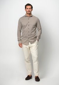 Camisa marrón claro de manga larga con botones oscuros, combinada con pantalones color crema y mocasines marrones, exhibida en un modelo contra un fondo neutro.