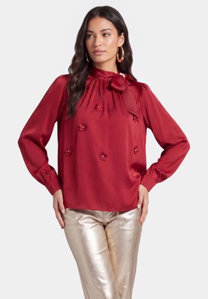 Kvinde iført en rød satinbluse med blomsterdetaljer og sløjfe ved halsen, kombineret med metallic guld bukser, stående med hænderne på hoften.