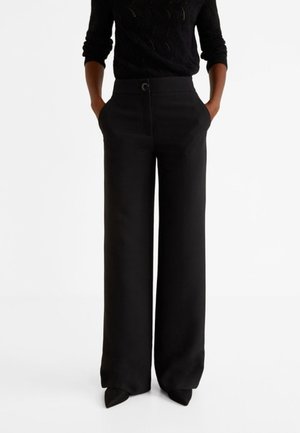 Osoba ubrana w czarne spodnie z wysokim stanem i szerokimi nogawkami z przodu z guzikami, ręce w kieszeniach, czarny długi rękaw i czarne szpiczaste buty.