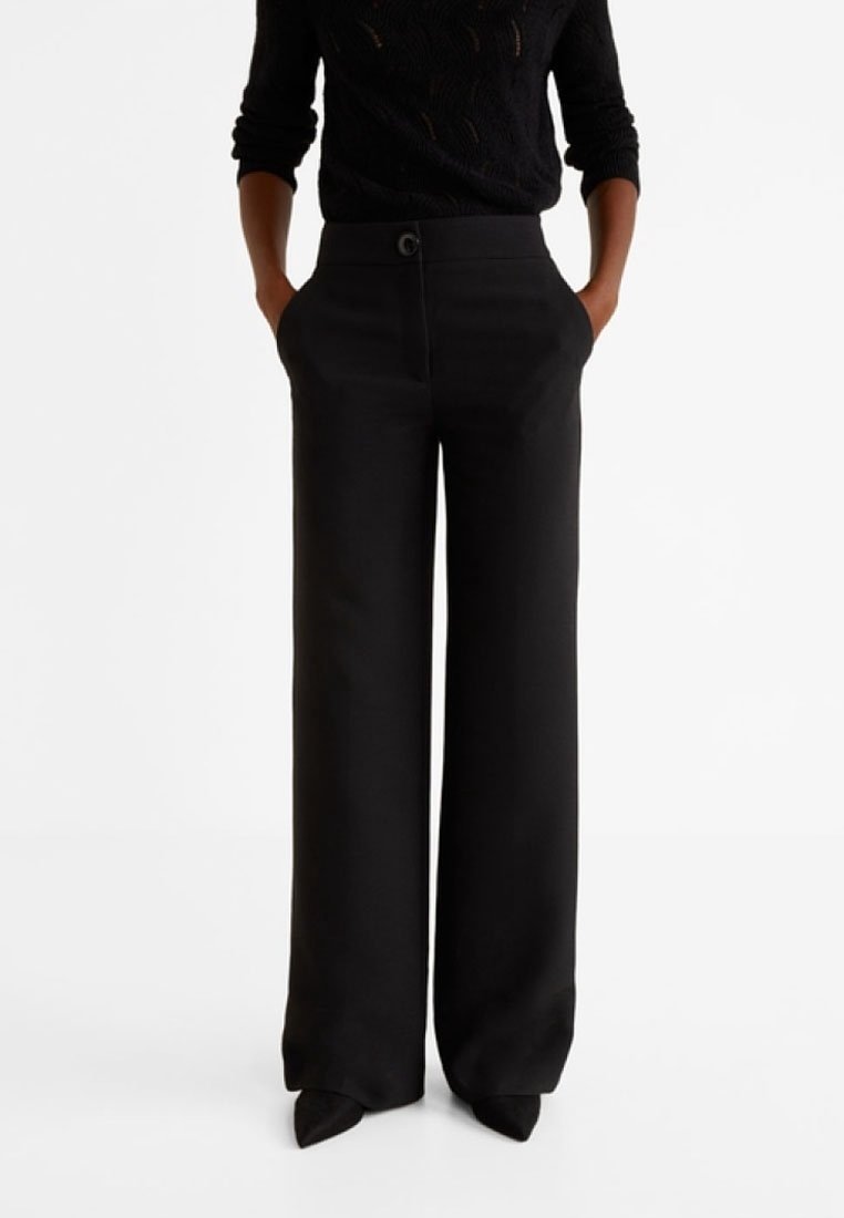 Personne portant un pantalon noir taille haute à jambes larges avec un bouton à l'avant, les mains dans les poches, un haut noir à manches longues et des chaussures pointues noires.
