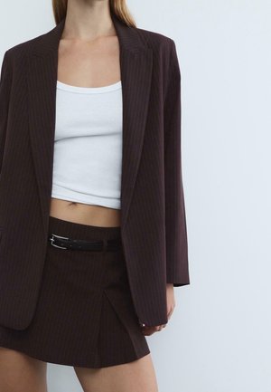Femme portant un blazer à fines rayures marron foncé et une jupe assortie avec une ceinture noire, associé à un débardeur court blanc.