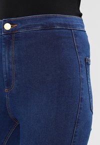 Jeans de mezclilla azul oscuro con una cintura alta, costuras visibles y un bolsillo frontal. El material parece suave y tiene un ligero estiramiento.