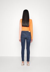 Mujer con cabello negro largo, vistiendo un suéter de bloques de color naranja y rosa, jeans ajustados oscuros y sandalias de tacón blanco, de pie mirando hacia fuera.