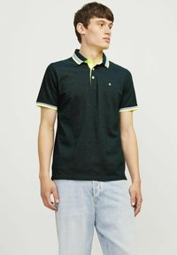 Polo shirt vert foncé avec un plastron à deux boutons, un col et des poignets avec des accents jaunes, et un petit logo sur la poitrine. Coupe décontractée, tissu texturé.