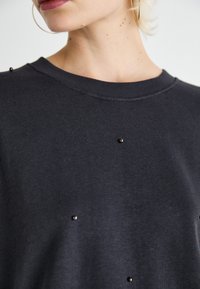 Mørk grå hettegenser med ribbestrikket hals, prydet med dekorative svarte perler jevnt fordelt over stoffet, som gir et teksturert utseende.