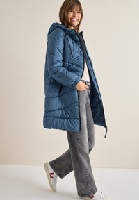 Piumino blu con cappuccio, caratterizzato da un design trapuntato e polsini elastici. Indossato sopra jeans grigi chiari e sneakers bianche con accenti rossi.