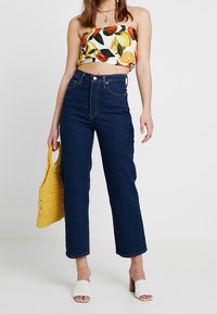 Jeans a vita alta, dritti in denim scuro, abbinati a un top a fiori corto. Gli accessori includono una borsa in crochet gialla e mule bianche con tacco.