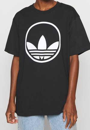 Henkilö, jolla on musta t-paita, jossa on suuri valkoinen pyöreä Adidas-logo, ja vaaleansiniset farkut.