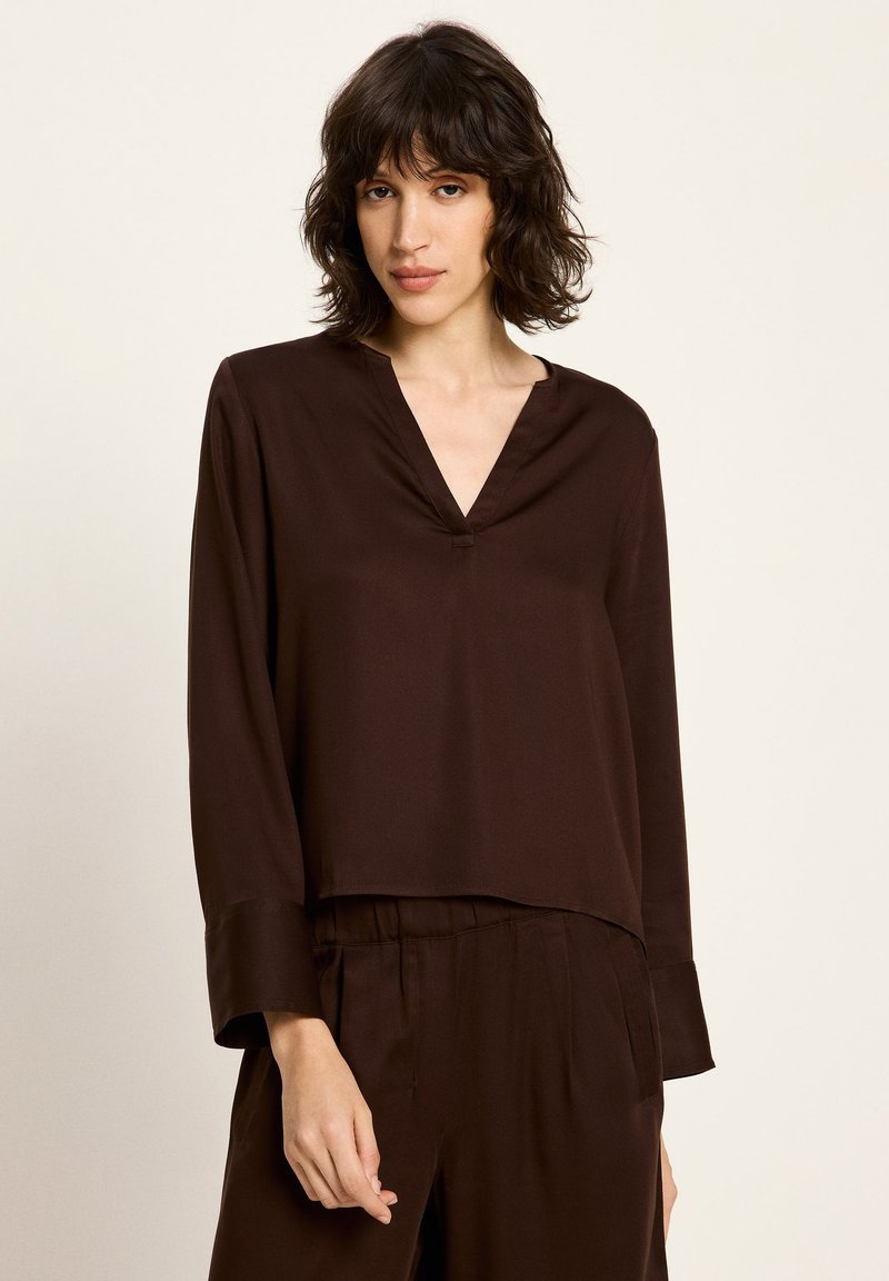 Frau mit schulterlangem welligem dunklem Haar, die eine dunkelbraune langärmelige V-Ausschnitt-Bluse und passende hochtaillierte Hosen vor einem schlichten Hintergrund trägt.