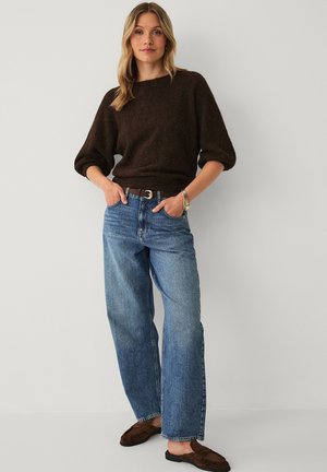 Vrouw draagt een donkerbruine trui, blauwe wijdere broek met een riem, bruine instapschoenen, staat met handen in de zakken tegen een lichte achtergrond.