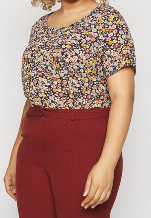 Vrouw draagt een kortemouws bloemenblouse met gele, oranje en witte bloemen, gecombineerd met hooggetailleerde donkerrode broek.