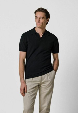 Polo nera a maniche corte con scollo a V, realizzata in tessuto liscio, abbinata a pantaloni sartoriali beige chiaro.