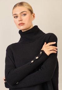 Czarny sweter w serek wykonany z prążkowanego dzianiny, o luźnym kroju, z długimi rękawami i ozdobnymi metalowymi guzikami przy mankietach.