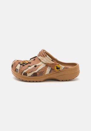 Sabot marron à enfiler avec imprimé animal beige et marron foncé, trous de ventilation et une bride avec un logo circulaire jaune sur fond blanc.