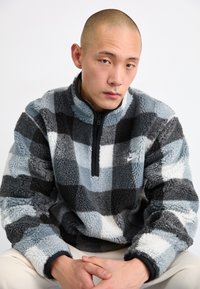 Szürke és fekete kockás mintájú fleece pulóver, magas nyakkal és félig zipzárazható. A mellkason egy kis Nike logó található.
