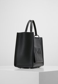 Czarna skórzana torba typu tote z podwójnymi uchwytami, teksturowaną przednią częścią i minimalistycznym designem. Wyposażona w metalowe nóżki dla stabilności.