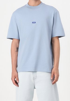 Homme portant un t-shirt à manches courtes bleu clair avec un petit logo "HUGO" bleu sur la poitrine et un jean bleu clair, debout devant un fond uni.