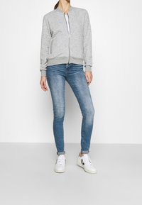 Grijze bomberjack met ritssluiting en gestructureerde stof, geribbelde manchetten, gecombineerd met lichtblauwe skinny jeans en witte sneakers met zwarte accenten.