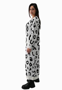 Langes Strickkleid in Weiß mit schwarzem Tierdruck. Hat eine lockere Passform, breite Ärmel, gerippte Bündchen und Taschen. Kombiniert mit schwarzen Stiefeln.