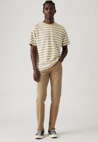 T-shirt en coton rayé beige et crème, avec un col rond et des manches courtes. Associé à un jean droit marron clair et des baskets noires.