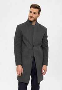 Grauer Wollmantel mit hohem Kragen und Ein-Knopf-Verschluss. Gerader Schnitt, strukturierter Stoff und lange Ärmel. Über einem blauen Hemd getragen.