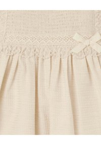 Abito beige chiaro con corpetto arricciato e bordo in pizzo, gonna plissettata e piccolo fiocco decorativo di lato. Tessuto texture.