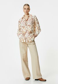 Button-up shirt in cream with abstract rust and brown geometric patterns; paired with beige wide-leg trousers and brown loafers.