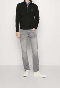 Man som bär en svart zip-tröja över en blå mönstrad skjorta, grå jeans och vita sneakers, står med en hand i fickan.