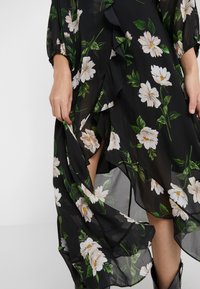 Zwart bloemenjurk van lichtgewicht chiffon, met witte bloemen en groene bladeren, voorzien van ruches langs de randen en mouwen.