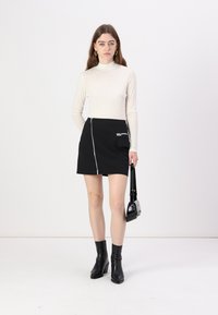 Gonna nera con zip e tasca laterale, abbinata a un top aderente a maniche lunghe color crema. La modella tiene in mano una piccola borsa nera.
