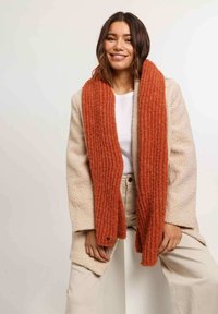 Abrigo de mezcla de lana beige con una textura suave, superpuesto a una blusa blanca. Presenta una bufanda de canalé naranja brillante, que añade contraste. Fondo neutro claro.