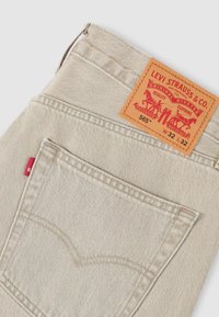 Μπεζ τζιν από denim της Levi's με δερμάτινο έμπλαστρο στη μέση και κόκκινη ταμπελίτσα στην πίσω τσέπη, με λεπτομέρειες ραφής σε αντίθετο χρώμα.