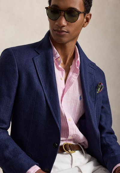 Mees, kellel on seljas tumesinine bleiser, roosas triibuline särk, valged püksid, pruun vöö, värviline taskurätik ja ümmargused päikeseprillid neutraalsel taustal.