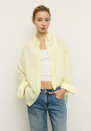 Kobieta z rękami w kieszeniach, ubrana w jasnożółtą koszulę zapinaną na guziki na białym topie odsłaniającym brzuch i niebieskie dżinsy, stojąca na tle jednolitej ściany.