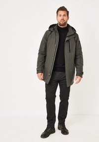 Grey insulated jacket with a hood, featuring a front zipper and button closure, black turtleneck, black pants, and dark boots.