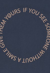 Navy fabric with circular text in light tan reading, "GIVE THEM YOURS IF YOU SEE SOMEONE WITHOUT A SMILE." Featuring a smiley face.